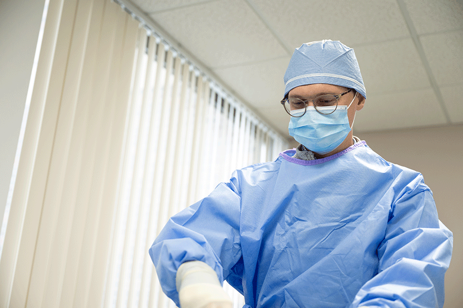 A white male physician wearing a blue surgical gown, a blue surgical mask, white surgical gloves, and a blue surgical cap. He also has dark brown glasses. He is looking down at his hands in a room with windows and window blinds visible behind him.   